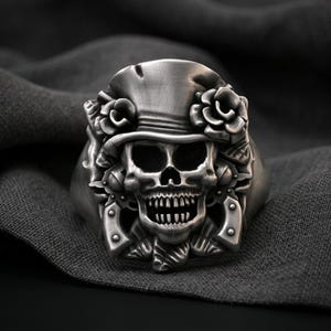 May include: A silver-toned metal ring featuring a detailed skull wearing a top hat adorned with roses. Two revolvers are positioned on either side of the skull, integrated into the band design with a brushed metallic finish and dark accents.