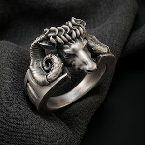 May include: A silver-toned ring featuring a detailed ram head sculpture as the centerpiece. The design includes prominent, textured curved horns that wrap around the sides of the band, which has a smooth, polished finish and a solid, thick construction.