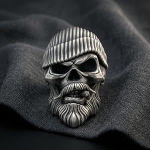 Op de afbeelding: Een zilverkleurige ring met een gedetailleerd schedelontwerp. De schedel draagt een geribbelde muts en heeft een opvallende snor en baard. Een klein, cilindervormig object dat op een sigaar lijkt, wordt tussen de tanden van de schedel gehouden.