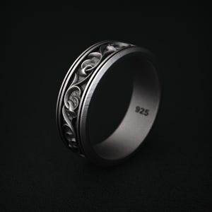 May include: A silver ring with an intricate floral pattern. The ring has a wide band with a detailed design of swirling leaves and vines. The metal is a matte silver finish, and the ring is marked with "925".
