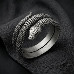 Puede incluir: Un brazalete de metal plateado con forma de serpiente enroscada. La superficie exterior presenta un patrón de escamas detallado y repetitivo, mientras que la cabeza de la serpiente se muestra claramente en un extremo. La banda interior tiene un acabado cepillado liso.