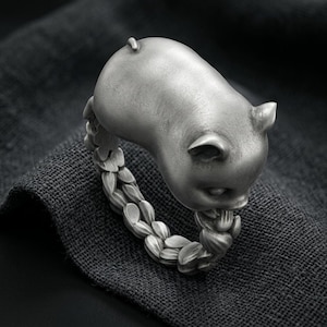 Puede incluir: Un anillo de tono plateado que presenta una figura de cerdo esculpida como pieza central. La banda del anillo está construida a partir de una serie de segmentos interconectados en forma de pétalos u hojas que se envuelven para formar la pieza.
