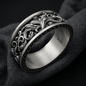 Op de afbeelding: Een brede zilveren ring met een ingewikkeld, verhoogd bloemrijk krulpatroon over de buitenste band. Het binnenoppervlak is glad met een geborstelde afwerking en toont duidelijk de gestempelde tekst 925, wat duidt op sterling zilver.
