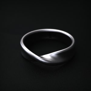 May include: A silver-colored ring with a continuous, twisted design. The ring has a smooth, polished surface and a minimalist aesthetic. The ring is shown against a dark background.