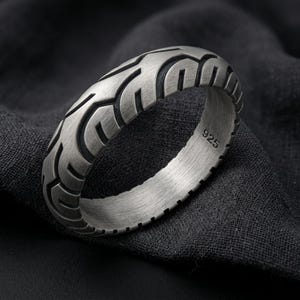 May include: A brushed silver-tone ring featuring a textured tread pattern reminiscent of a tire. The outer surface displays deep, carved grooves in a repeating design, while the inner band is clearly stamped with the number 925 for authenticity.