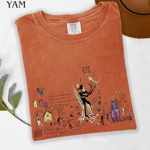 Puede incluir: Una camiseta de cuello redondo color naranja quemado con una etiqueta Comfort Colors y un estampado gr&aacute;fico complejo. El dise&ntilde;o incluye una figura negra central, peque&ntilde;as casas coloridas, varios bocetos abstractos y texto escrito a mano que incluye la palabra UNREALIZED.