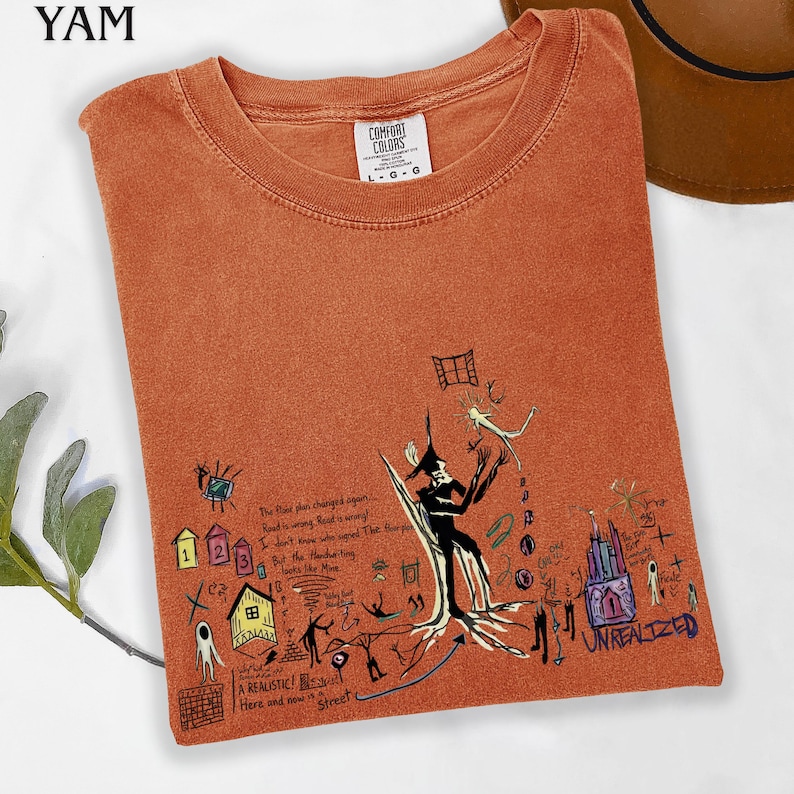 Puede incluir: Una camiseta de cuello redondo color naranja quemado con una etiqueta Comfort Colors y un estampado gr&aacute;fico complejo. El dise&ntilde;o incluye una figura negra central, peque&ntilde;as casas coloridas, varios bocetos abstractos y texto escrito a mano que incluye la palabra UNREALIZED.