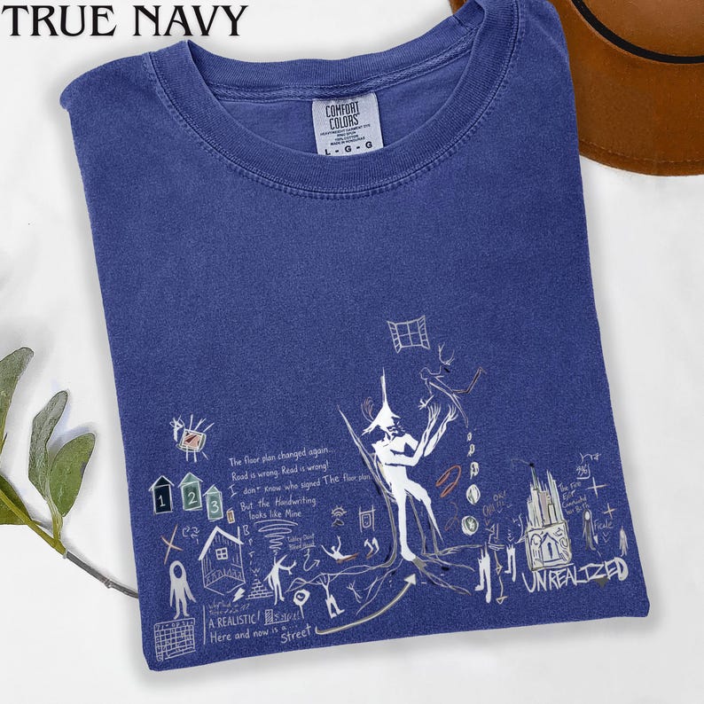 Puede incluir: Una camiseta azul marino con un estampado gr&aacute;fico blanco que incluye varios bocetos dibujados a mano, como una figura central, casas peque&ntilde;as, un edificio y el texto The floor plan changed again, Road is wrong y UNREALIZED.