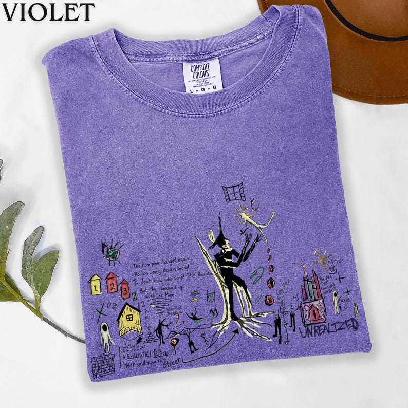Puede incluir: Una camiseta de cuello redondo color violeta con un estampado gr&aacute;fico negro que incluye varias ilustraciones dibujadas a mano, como casas, una figura central y el texto "The floor plan changed again" y "UNREALIZED" en estilo boceto.