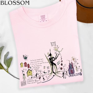 Puede incluir: Una camiseta rosa claro con un gr&aacute;fico complejo de estilo dibujo a mano en negro, rojo, amarillo y azul. El dise&ntilde;o incluye varias ilustraciones peque&ntilde;as, figuras abstractas y texto escrito a mano como "The floor plan changed again" y "UNREALIZED".