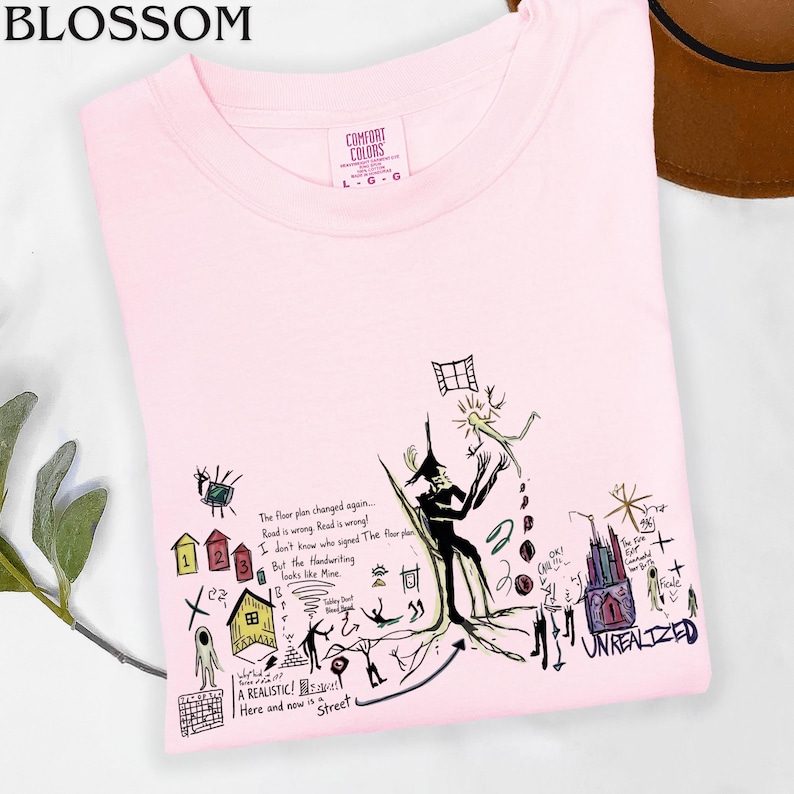 Puede incluir: Una camiseta rosa claro con un gr&aacute;fico complejo de estilo dibujo a mano en negro, rojo, amarillo y azul. El dise&ntilde;o incluye varias ilustraciones peque&ntilde;as, figuras abstractas y texto escrito a mano como "The floor plan changed again" y "UNREALIZED".
