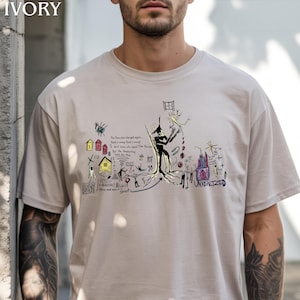 Puede incluir: Una camiseta de color marfil de manga corta con un complejo estampado gr&aacute;fico multicolor. El dise&ntilde;o incluye varios bocetos dibujados a mano, figuras abstractas, peque&ntilde;as casas numeradas 1, 2, 3 y texto escrito a mano, incluyendo UNREALIZED.