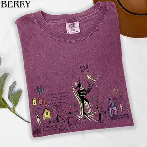 Puede incluir: Una camiseta de cuello redondo color baya con una etiqueta Comfort Colors y un estampado gr&aacute;fico oscuro. El dise&ntilde;o incluye varias ilustraciones de estilo dibujo a mano, como una figura central, casas peque&ntilde;as y el texto "UNREALIZED" y "A REALISTIC".
