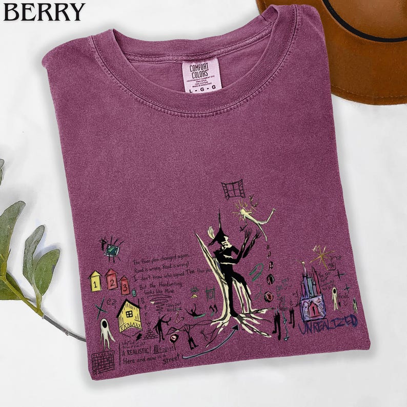 Puede incluir: Una camiseta de cuello redondo color baya con una etiqueta Comfort Colors y un estampado gr&aacute;fico oscuro. El dise&ntilde;o incluye varias ilustraciones de estilo dibujo a mano, como una figura central, casas peque&ntilde;as y el texto "UNREALIZED" y "A REALISTIC".