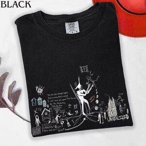 Puede incluir: Una camiseta negra de manga corta con un estampado gr&aacute;fico blanco que incluye varias ilustraciones dibujadas a mano, como una figura central, iconos de casas peque&ntilde;as y el texto The floor plan changed again y UNREALIZED.