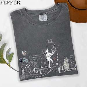 Puede incluir: Una camiseta de cuello redondo gris carb&oacute;n con una etiqueta Comfort Colors en el cuello. El frente presenta un estampado gr&aacute;fico blanco de una figura central, casas peque&ntilde;as, una ventana y texto escrito a mano que dice "The floor plus changed again..." y "UNREALIZED".
