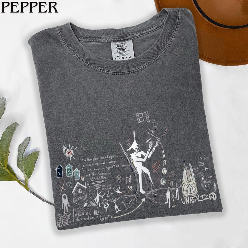 Puede incluir: Una camiseta de cuello redondo gris carb&oacute;n con una etiqueta Comfort Colors en el cuello. El frente presenta un estampado gr&aacute;fico blanco de una figura central, casas peque&ntilde;as, una ventana y texto escrito a mano que dice "The floor plus changed again..." y "UNREALIZED".