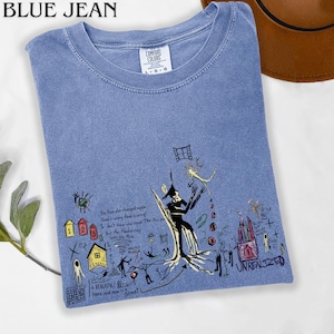 Puede incluir: Una camiseta azul de cuello redondo con etiqueta Comfort Colors, que presenta un estampado gr&aacute;fico de dibujos surrealistas en l&iacute;neas negras, peque&ntilde;as casas coloridas y el texto "UNREALIZED" junto a varias notas manuscritas y bocetos abstractos.