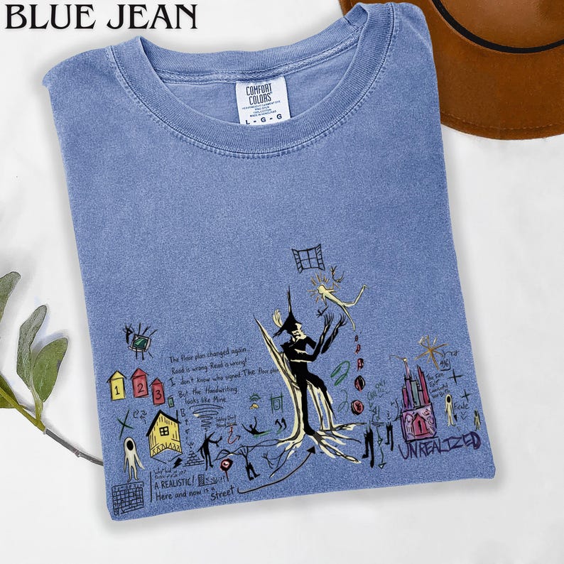 Puede incluir: Una camiseta azul de cuello redondo con etiqueta Comfort Colors, que presenta un estampado gr&aacute;fico de dibujos surrealistas en l&iacute;neas negras, peque&ntilde;as casas coloridas y el texto "UNREALIZED" junto a varias notas manuscritas y bocetos abstractos.