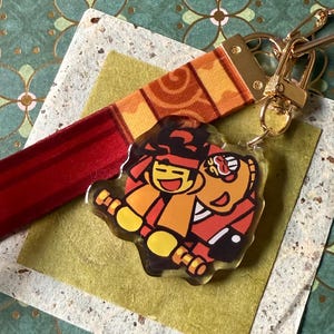 May include: A keychain featuring a fabric strap with a red and orange pattern and a gold-toned metal clasp. Attached is a clear acrylic charm displaying a colorful cartoon illustration of two figures, one with a red headband, against a red circle.