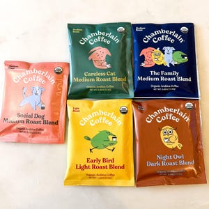 Puede incluir: Seis paquetes coloridos de café Chamberlain, cada uno con una ilustración de animal diferente. Los paquetes están etiquetados con tipos de tueste como "Medium" y "Dark", y presentan texto como "Careless Cat" y "Early Bird".