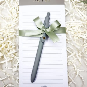 May include: A notepad with the word "NOTES" in gold lettering, a gray pen, and a sage green ribbon. The notepad has lined paper and a small floral design at the bottom. The pen is secured to the notepad with the ribbon.