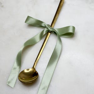 以下が含まれることがあります： 薄緑色のサテンリボンで結ばれた金色のスプーン。スプーンは小さく丸いボウルと長く細い柄をしています。リボンは蝶結びになっており、長く流れるような端があります。スプーンとリボンは白い大理石の表面にあります。