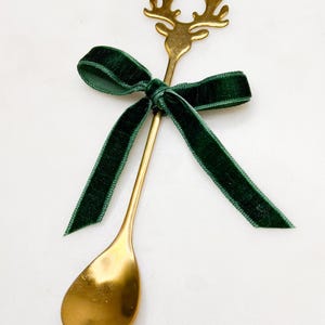May include: A gold-coloured spoon featuring a reindeer head design and a dark green velvet ribbon tied around the handle. The spoon has a rounded bowl and is set against a white background. A festive, decorative item.