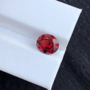 May include: A faceted, oval-shaped, deep red gemstone. The gem is set against a white surface, with a dark background. The gemstone's cut allows light to reflect, creating a brilliant appearance. This gemstone could be used in jewelry.