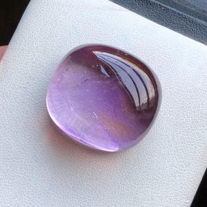 Könnte beinhalten: Ein polierter, abgerundeter Amethyst-Edelstein mit einem Farbverlauf von tiefem Lila zu durchscheinendem Lavendel. Der Stein hat eine glatte, reflektierende Oberfläche und ist vor einem weißen Hintergrund platziert.