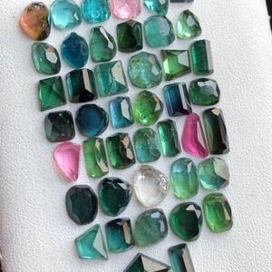 May include: A selection of cut gemstones in diverse shapes and hues, such as teal, pink, green, and clear. The gemstones are displayed on a white surface, highlighting their varied cuts and clarity.