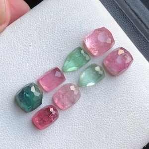 May include: A collection of faceted gemstones in various colors, including pink, green, and teal. The gems are displayed on a white surface, showcasing their polished surfaces and unique shapes. The gemstones are likely for jewelry making.