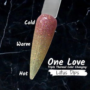 May include: A nail swatch with a color-changing effect. The top is pink with glitter, transitioning to gold at the bottom. The text reads "One Love Triple Thermal Color Changing Lotus Dips."