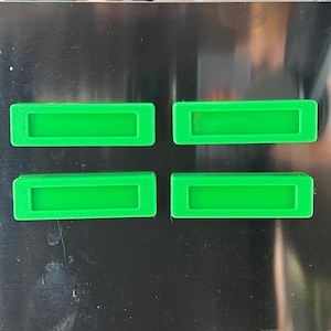 May include: Four bright green rectangular objects with a recessed center are arranged on a dark surface. The objects appear to be made of plastic or a similar material, and are evenly spaced.
