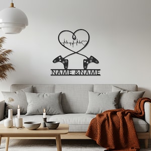 Puede incluir: Arte de pared de metal negro con un diseño en forma de corazón con una línea de latidos y dos mandos de juego. El texto "NAME & NAME" se muestra debajo. El arte está montado en una pared blanca sobre un sofá gris.