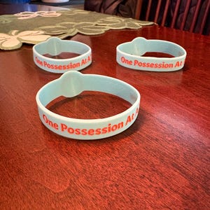 May include: Three light blue silicone wristbands with the text "One Possession At A" printed in red. The wristbands are on a dark red wooden surface. The wristbands have a raised, rounded section.