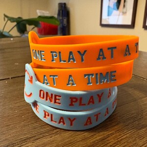 Puede incluir: Pila de pulseras de silicona naranja y azul claro. Las bandas naranjas tienen el texto "ONE PLAY AT A TIME" en letras plateadas. Las bandas azul claro tienen el mismo texto en letras rojas.