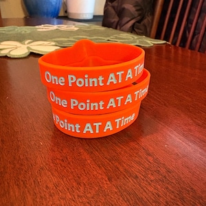 May include: A stack of three orange silicone wristbands. Each band features the text "One Point AT A Time" in light blue lettering. The bands are stacked on a dark wooden surface.