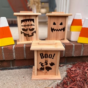 May include: Wooden Halloween-themed boxes with carved designs of a Frankenstein monster, a jack-o'-lantern face, and a paw print with the word "BOO!". Painted candy corn decorations are also present, adding to the festive atmosphere.
