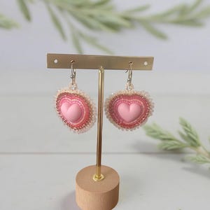 May include: Pink heart-shaped earrings with a beaded border and a smaller heart in the centre, displayed on a gold stand with a wooden base. The earrings have a handcrafted appearance. The earrings are a charming accessory.