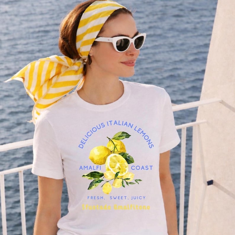 May include: White t-shirt with a watercolour illustration of lemons and the text "Delicious Italian Lemons Amalfi Coast". The shirt also has the text "Fresh, Sweet, Juicy". The person is wearing a yellow and white striped scarf and white sunglasses.