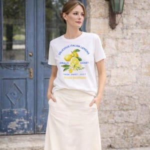 May include: A white t-shirt with a lemon graphic and the text "Delicious Italian Lemons Amalfi Coast". The shirt is worn with a cream skirt. The background shows a blue door and a stone wall.