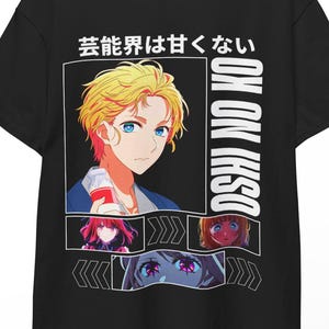 Może przedstawiać: Czarny t-shirt z grafiką w stylu anime. Projekt zawiera męską postać z blond włosami i niebieskimi oczami, japoński tekst i dodatkowe portrety postaci. Tekst "OSHI NO KO" jest wydrukowany pionowo po prawej stronie.