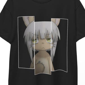 Puede incluir: Camiseta negra con un personaje de dibujos animados de pelo blanco, orejas de conejo y ojos verdes, enmarcado en un diseño rectangular blanco. El personaje está centrado en la camiseta.