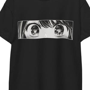 Może przedstawiać: Czarny t-shirt z czarno-białą ilustracją w stylu mangi przedstawiającą oczy. Prostokątna grafika jest wyśrodkowana na koszulce. Koszulka wisi na drewnianym wieszaku.