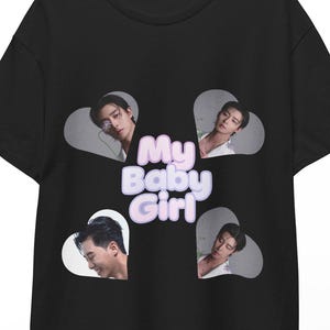 May include: Black t-shirt featuring four images of a person within white heart shapes. The text "My Baby Girl" is printed in a light purple color on the front of the shirt. The shirt is hanging on a wooden hanger.