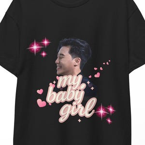 May include: Black t-shirt featuring a graphic of a smiling person with the text "my baby girl" in a cursive font. Pink hearts and starbursts surround the image. The t-shirt is hanging on a wooden hanger.