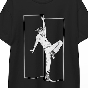 Puede incluir: Camiseta negra con una ilustración en blanco y negro de una persona en una pose dinámica, enmarcada por un rectángulo blanco. La persona lleva gafas, una camisa de manga larga y pantalones. La camiseta está colgada en una percha de madera.