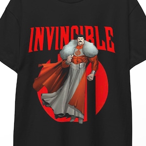 May include: Black t-shirt featuring a graphic of a character in a red and white costume with a red cape. The word "INVINCIBLE" is printed in red above the character. The character is standing in front of a red circle.