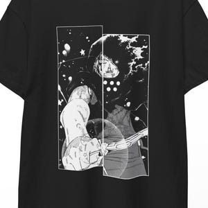 Puede incluir: Camiseta negra con un gráfico en blanco y negro estilo manga. El gráfico representa una figura con una capa con capucha y un arma. El diseño está dividido en dos paneles verticales. La camiseta está colgada en una percha de madera.